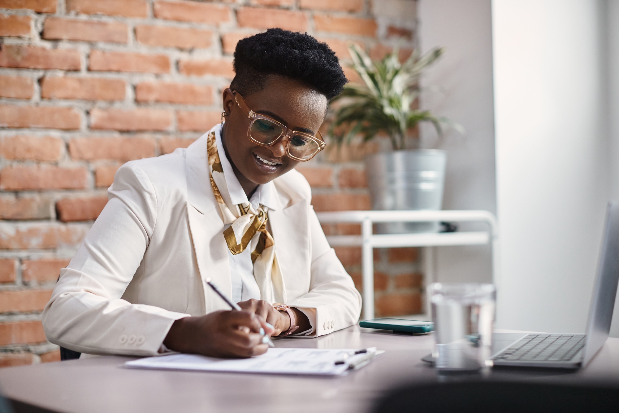 Happy black businesswoman writing report while working on laptop at corporate office.